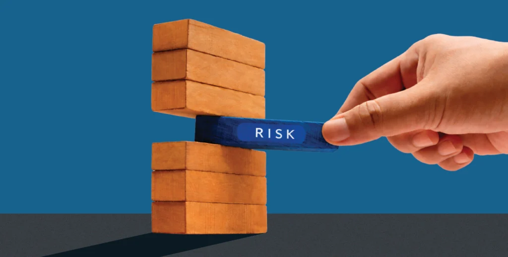 Hand removing a blue wooden block labeled “Risk” from a stacked wooden tower, symbolizing risk management and structural stability in financial systems.