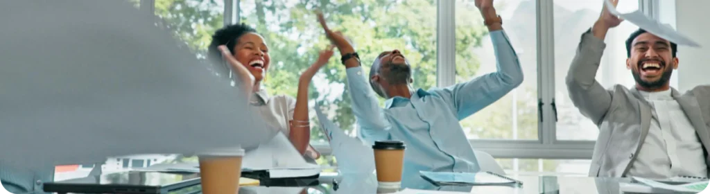 Three diverse colleagues laughing and celebrating at a conference table, throwing papers in the air in a bright, modern office with large windows and greenery in the background