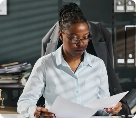 A smiling professional woman with braided hair wearing a blazer, engaged in a conversation at a desk with a desk phone visible in the foreground, in a busy office setting